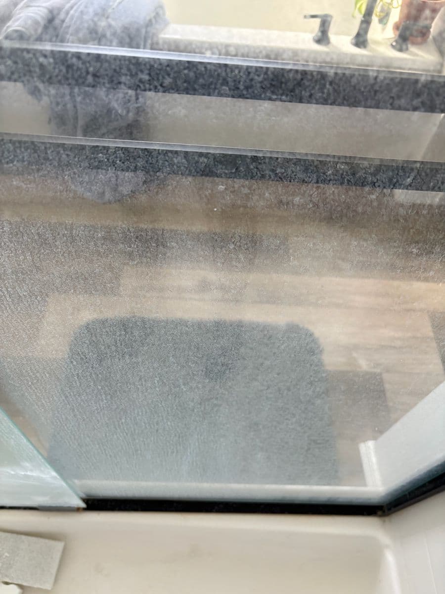Cloudy shower glass before cleaning, bath mat and wood floor barely visible through hard-water haze