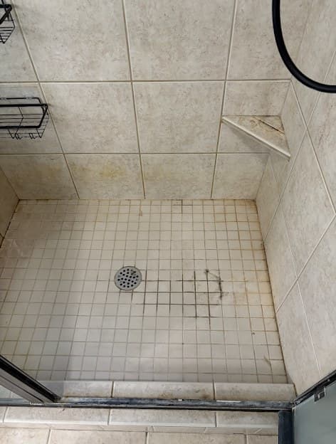 Walk-in shower before cleaning, beige tile floor and grout stained with dark grime and hard-water residue around the drain